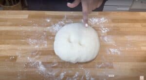 Finger pressing dough ball on floured wooden surface.